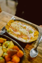 A metal oval dish contains a colorful meal with chunks of green avocado, cooked chickpeas, shredded cheese, and plantain chips, topped with a creamy sauce and garnished with herbs. Another dish filled with various cooked vegetables, such as potatoes and sweet potatoes, is partially visible in the foreground. Both dishes rest on a rustic wooden table beside a spoon.