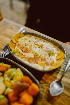 A metal oval dish contains a colorful meal with chunks of green avocado, cooked chickpeas, shredded cheese, and plantain chips, topped with a creamy sauce and garnished with herbs. Another dish filled with various cooked vegetables, such as potatoes and sweet potatoes, is partially visible in the foreground. Both dishes rest on a rustic wooden table beside a spoon.