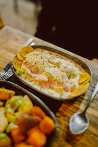 A metal oval dish contains a colorful meal with chunks of green avocado, cooked chickpeas, shredded cheese, and plantain chips, topped with a creamy sauce and garnished with herbs. Another dish filled with various cooked vegetables, such as potatoes and sweet potatoes, is partially visible in the foreground. Both dishes rest on a rustic wooden table beside a spoon.