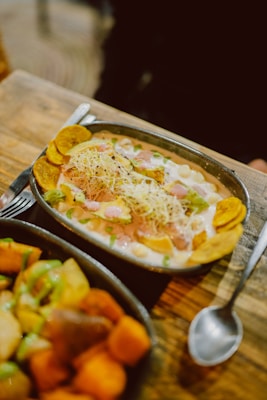 A metal oval dish contains a colorful meal with chunks of green avocado, cooked chickpeas, shredded cheese, and plantain chips, topped with a creamy sauce and garnished with herbs. Another dish filled with various cooked vegetables, such as potatoes and sweet potatoes, is partially visible in the foreground. Both dishes rest on a rustic wooden table beside a spoon.