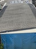 A large corrugated metal roof with a section of blue tarp covering the area at the bottom. There are some plants visible at the lower left corner.