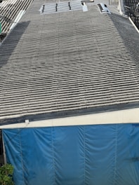 A large corrugated metal roof with a section of blue tarp covering the area at the bottom. There are some plants visible at the lower left corner.