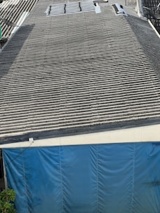 A large corrugated metal roof with a section of blue tarp covering the area at the bottom. There are some plants visible at the lower left corner.