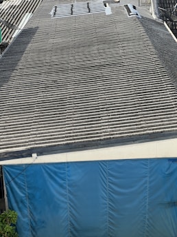 A large corrugated metal roof with a section of blue tarp covering the area at the bottom. There are some plants visible at the lower left corner.