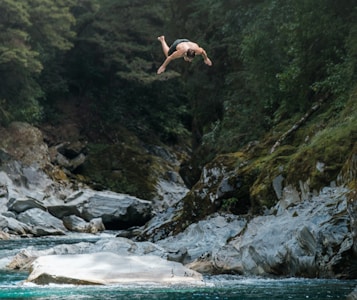 A person is captured in mid-air above a clear, flowing river surrounded by dense forest and large rocks. The individual appears to be performing a dive or jump, wearing swim trunks. The setting is natural and untouched, invoking a sense of adventure and freedom.