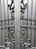 An ornate iron gate featuring intricate scrollwork designs. The symmetrical pattern includes elegant curves and twists, created from dark metal rods. The gate comprises vertical bars with decorative elements, providing a sense of both strength and artistic flair.