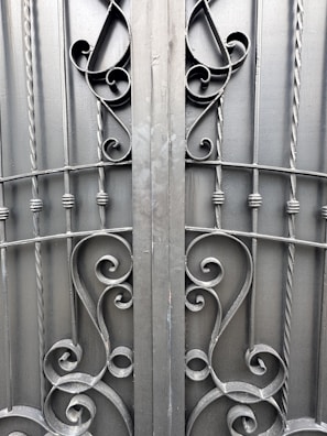 An ornate iron gate featuring intricate scrollwork designs. The symmetrical pattern includes elegant curves and twists, created from dark metal rods. The gate comprises vertical bars with decorative elements, providing a sense of both strength and artistic flair.