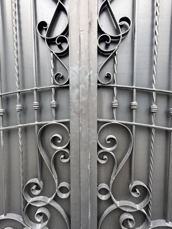 An ornate iron gate featuring intricate scrollwork designs. The symmetrical pattern includes elegant curves and twists, created from dark metal rods. The gate comprises vertical bars with decorative elements, providing a sense of both strength and artistic flair.