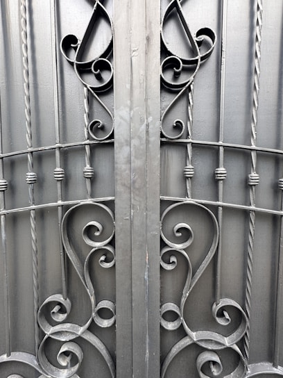 An ornate iron gate featuring intricate scrollwork designs. The symmetrical pattern includes elegant curves and twists, created from dark metal rods. The gate comprises vertical bars with decorative elements, providing a sense of both strength and artistic flair.
