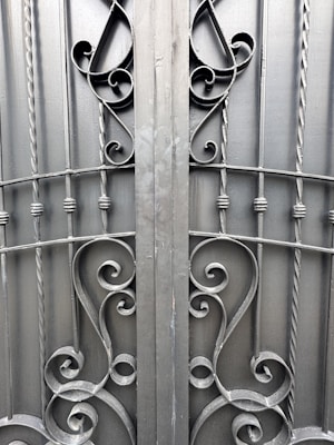 An ornate iron gate featuring intricate scrollwork designs. The symmetrical pattern includes elegant curves and twists, created from dark metal rods. The gate comprises vertical bars with decorative elements, providing a sense of both strength and artistic flair.