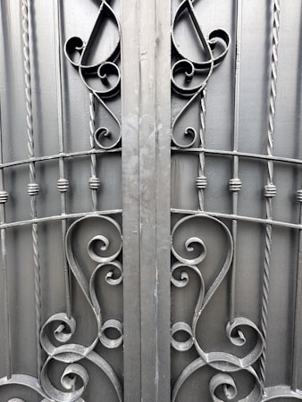 An ornate iron gate featuring intricate scrollwork designs. The symmetrical pattern includes elegant curves and twists, created from dark metal rods. The gate comprises vertical bars with decorative elements, providing a sense of both strength and artistic flair.