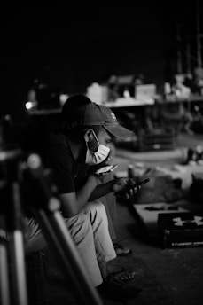 A person wearing a cap and a face mask is sitting and looking intently at a smartphone. The background is blurred, with objects like tables, boxes, and potentially other people visible. The setting appears to be indoors, possibly a workspace or studio environment with dim lighting.