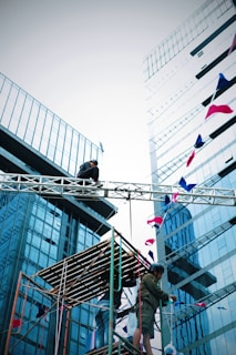 Workers are on a scaffolding structure surrounded by tall modern glass buildings. Flags are draped across a metal framework. The scene suggests construction or event setup.