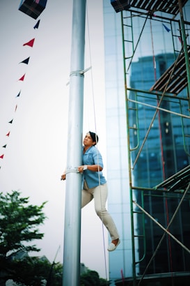 A person is climbing a tall metal pole, surrounded by a structure that resembles scaffolding. The background features a modern glass building with a clear sky overhead. Colorful triangular flags are strung along a line near the pole. The person appears focused, wearing a blue shirt and light pants.