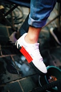 A person's foot wearing a white sneaker with a striking red and black strip on the sole. The person is wearing blue rolled-up jeans. The shoe is labeled with some text, placed above a wet pavement, next to some indistinguishable items and a green container.