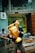 A person is holding a large golden trophy labeled 'PROMOSI' while looking upward. The setting seems to be outdoors near a modern building. In the background, another person is seated, engrossed in a mobile device, with construction materials visible nearby.