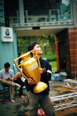 A person is holding a large golden trophy labeled 'PROMOSI' while looking upward. The setting seems to be outdoors near a modern building. In the background, another person is seated, engrossed in a mobile device, with construction materials visible nearby.
