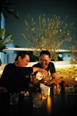 Two people are enjoying a lively moment together at a dimly lit table. They are sharing drinks, laughing, and surrounded by a warm, cozy atmosphere. The scene is set outdoors with olive branches in the background, suggesting a relaxed and intimate environment.