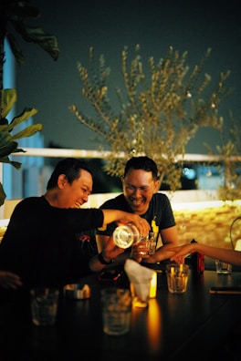 Two people are enjoying a lively moment together at a dimly lit table. They are sharing drinks, laughing, and surrounded by a warm, cozy atmosphere. The scene is set outdoors with olive branches in the background, suggesting a relaxed and intimate environment.