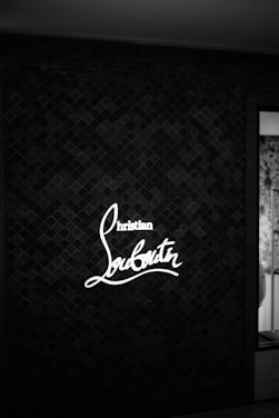 A monochrome image featuring an illuminated 'Christian Louboutin' sign against a textured wall with a scale-like pattern. The light contrasts with the dark background, making the text stand out.