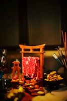 A wooden frame resembling a torii gate holds slices of marbled red meat. Nearby, various food items are artistically arranged on a table, including round treats, sliced vegetables, and a bowl of garnished dish. Chopsticks and other dining utensils are visible to the side.