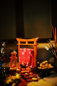 A wooden frame resembling a torii gate holds slices of marbled red meat. Nearby, various food items are artistically arranged on a table, including round treats, sliced vegetables, and a bowl of garnished dish. Chopsticks and other dining utensils are visible to the side.