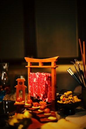 A wooden frame resembling a torii gate holds slices of marbled red meat. Nearby, various food items are artistically arranged on a table, including round treats, sliced vegetables, and a bowl of garnished dish. Chopsticks and other dining utensils are visible to the side.