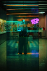 A dimly lit and reflective glass panel displays the word 'alchemist' in lowercase letters. Behind the glass, there appears to be a blurred interior with colorful lighting, including a pink neon sign. The scene is infused with a mix of green, blue, and purple hues, enhancing the moody atmosphere.