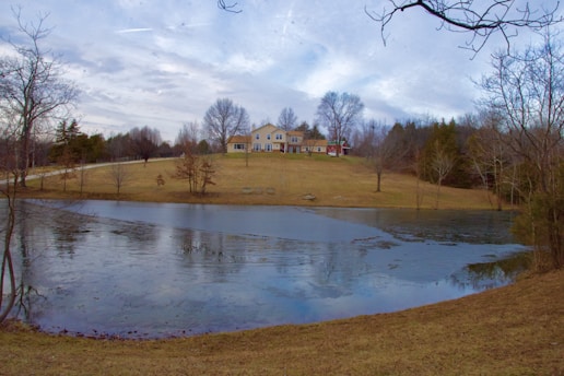 A large, elegant house stands on a gentle hill surrounded by sparse trees and a mown lawn. In the foreground, a partially frozen pond reflects the overcast sky. Bare branches and evergreens indicate a winter setting.
