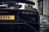 The rear view of a sleek and modern Lamborghini sports car, focusing on its distinctive taillights, aggressive bumper design, and a prominent exhaust system. The yellow license plate stands out against the dark exterior, and the car is parked on a patterned tile surface next to a building with large windows.