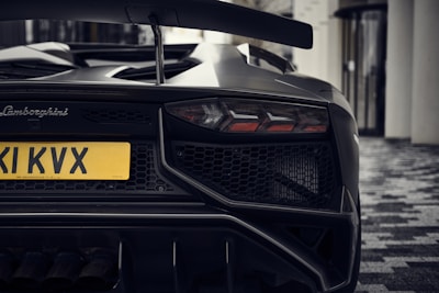 The rear view of a sleek and modern Lamborghini sports car, focusing on its distinctive taillights, aggressive bumper design, and a prominent exhaust system. The yellow license plate stands out against the dark exterior, and the car is parked on a patterned tile surface next to a building with large windows.