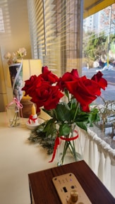 A bouquet of vibrant red roses is elegantly placed in a glass vase, adorned with green leaves and a red ribbon. The setup rests on a light-colored surface near a window with blinds, allowing natural light to filter in. Accompanying the roses are a decorative candle and a small bunch of white flowers in a vase nearby. Some pine branches add a touch of greenery, and a piece of wooden electronic equipment with a USB port is also visible.