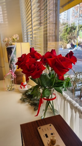 A bouquet of vibrant red roses is elegantly placed in a glass vase, adorned with green leaves and a red ribbon. The setup rests on a light-colored surface near a window with blinds, allowing natural light to filter in. Accompanying the roses are a decorative candle and a small bunch of white flowers in a vase nearby. Some pine branches add a touch of greenery, and a piece of wooden electronic equipment with a USB port is also visible.