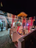 A two-story building decorated for a celebration with colorful drapes and strings of lights hanging across the courtyard. The building's windows and columns are illuminated, and there is a sign with the word 'BRID' illuminated below. The atmosphere is festive with a focus on vibrant lighting and decoration.