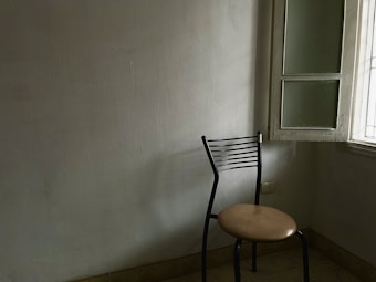 A simple room with an empty black metal chair placed near an open window. The walls are plain and the lighting is soft, streaming through the window to cast subtle shadows on the floor and wall.