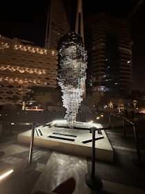 A unique sculpture composed of multiple horizontal layers, seemingly stacked to form an abstract human face, is displayed in an open area at night. Surrounding it are modern buildings with illuminated architectural designs. The sculpture is lit up from below, casting dramatic shadows and highlights.