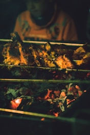 Grilled chicken pieces sitting above glowing coals on a barbecue. A person in the background is slightly blurred, wearing an orange garment.