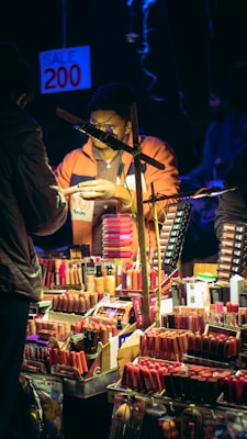 A vendor is selling a variety of cosmetics, including lipsticks and other makeup products, at a market stall. The display showcases numerous colorful cosmetic items neatly arranged. A sign above indicates a sale price of 200. The scene is dimly lit, with the warm glow focusing on the products and the vendor.