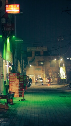 A dimly lit street at night with several illuminated signboards in Hindi, including one for a dhaba. The foreground features a green-tinted light on the pavement, while several vehicles and figures are visible in the background.