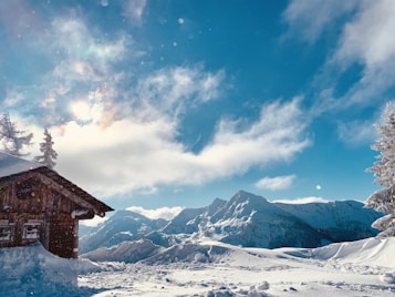 A snowy mountain landscape under a bright blue sky with scattered clouds. A wooden cabin is nestled in the foreground, partly covered in snow, while a pine tree stands nearby. The sun shines through the clouds, creating a serene and peaceful atmosphere.