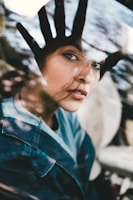 A woman looks through a glass window with her hand placed on it, creating an impression of separation or longing. Her expression is contemplative, with loose strands of hair gently brushing her face. The reflection on the glass and the soft lighting add a dreamy quality to the image.