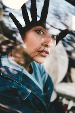 A woman looks through a glass window with her hand placed on it, creating an impression of separation or longing. Her expression is contemplative, with loose strands of hair gently brushing her face. The reflection on the glass and the soft lighting add a dreamy quality to the image.