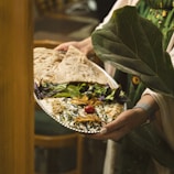 A person is holding a silver platter adorned with an intricately presented dish. The dish features a creamy base topped with a cherry tomato, drizzled with dark sauce, and garnished with herbs and shredded vegetables. Accompanying the dish are fresh greens and large flatbreads. The setting is cozy and rustic, with hints of wooden furniture in the background.