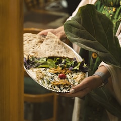 A person is holding a silver platter adorned with an intricately presented dish. The dish features a creamy base topped with a cherry tomato, drizzled with dark sauce, and garnished with herbs and shredded vegetables. Accompanying the dish are fresh greens and large flatbreads. The setting is cozy and rustic, with hints of wooden furniture in the background.