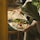 A person is holding a silver platter adorned with an intricately presented dish. The dish features a creamy base topped with a cherry tomato, drizzled with dark sauce, and garnished with herbs and shredded vegetables. Accompanying the dish are fresh greens and large flatbreads. The setting is cozy and rustic, with hints of wooden furniture in the background.