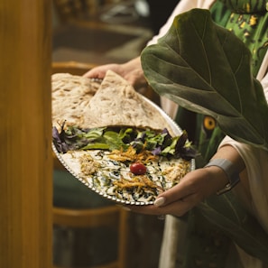 A person is holding a silver platter adorned with an intricately presented dish. The dish features a creamy base topped with a cherry tomato, drizzled with dark sauce, and garnished with herbs and shredded vegetables. Accompanying the dish are fresh greens and large flatbreads. The setting is cozy and rustic, with hints of wooden furniture in the background.