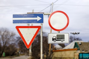 A collection of road signs including a blue sign with a white arrow pointing right, a yield sign with a red triangle border and a white center, a round red-bordered sign, and a rectangular sign depicting a truck. These signs are mounted on metal poles with a clear blue sky and trees in the background.