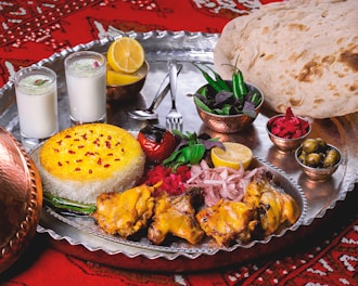 A lavish spread of Middle Eastern or South Asian cuisine is presented on a large metal tray. There are servings of yellow saffron rice and grilled chicken with a side of onions, garnished with lemon and fresh herbs. Two glasses of a yogurt-based drink, possibly lassi, are seen alongside. Additional side dishes include a bowl of green chili peppers and herbs, pickled vegetables, and a small bowl of olives. A large flatbread rests on the side, and the scene is set against a red patterned fabric.