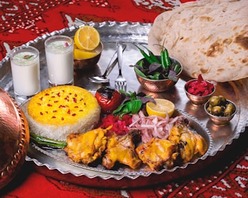 A lavish spread of Middle Eastern or South Asian cuisine is presented on a large metal tray. There are servings of yellow saffron rice and grilled chicken with a side of onions, garnished with lemon and fresh herbs. Two glasses of a yogurt-based drink, possibly lassi, are seen alongside. Additional side dishes include a bowl of green chili peppers and herbs, pickled vegetables, and a small bowl of olives. A large flatbread rests on the side, and the scene is set against a red patterned fabric.