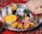 A lavish spread of Middle Eastern or South Asian cuisine is presented on a large metal tray. There are servings of yellow saffron rice and grilled chicken with a side of onions, garnished with lemon and fresh herbs. Two glasses of a yogurt-based drink, possibly lassi, are seen alongside. Additional side dishes include a bowl of green chili peppers and herbs, pickled vegetables, and a small bowl of olives. A large flatbread rests on the side, and the scene is set against a red patterned fabric.